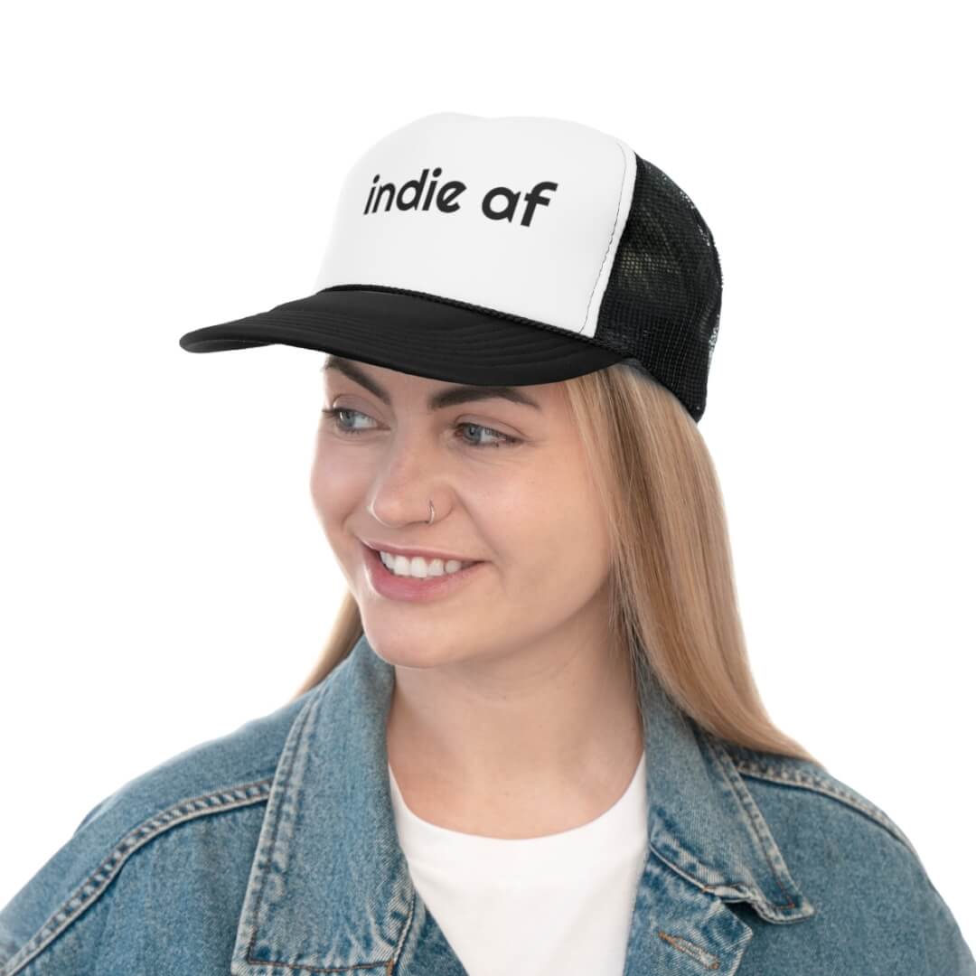 A woman wears the Indie AF Trucker Hat and a denim jacket, smiling while looking slightly to the side for a cool indie vibe.