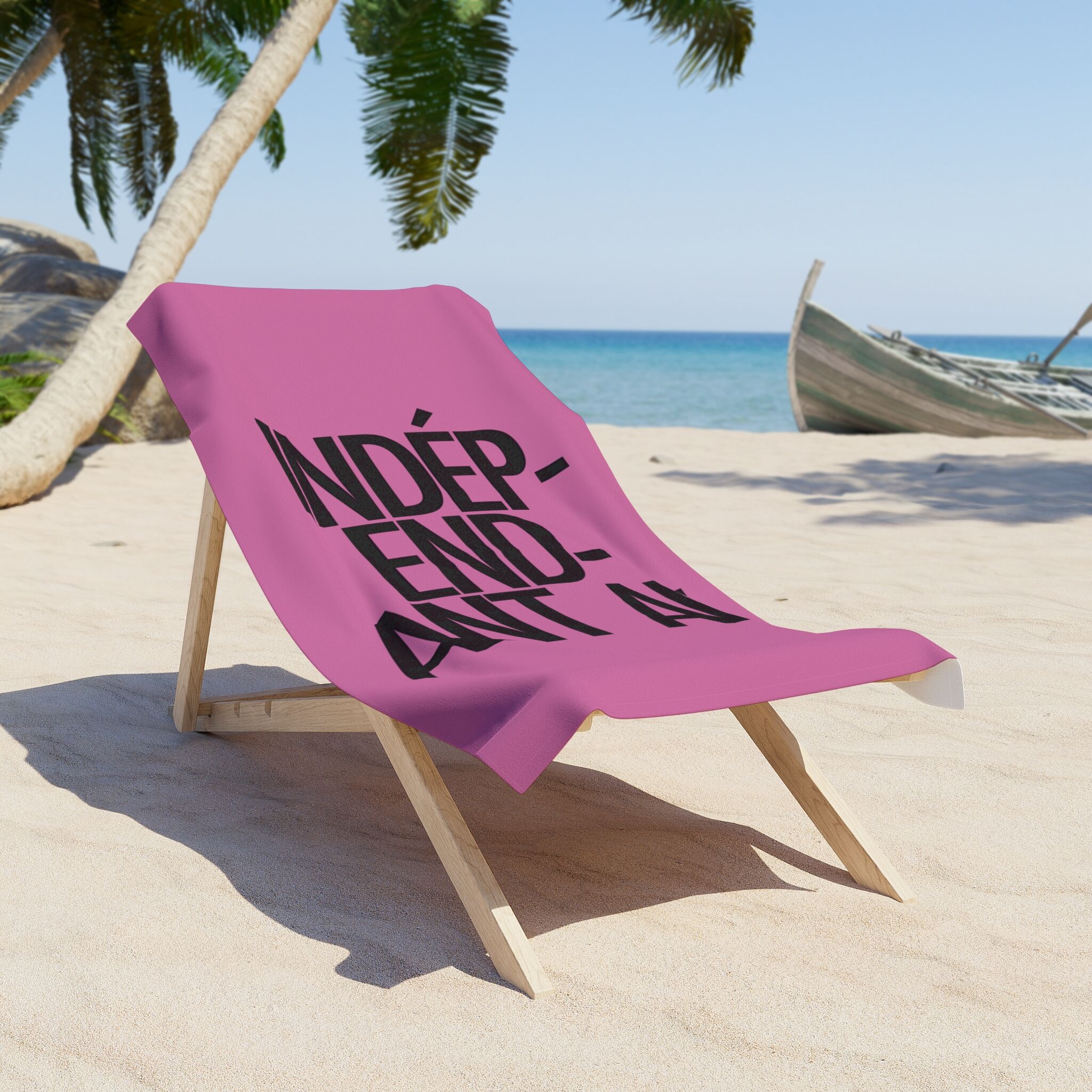 The Catherine Deneuve Would Approve Beach Towel—a pink towel with INDÉPENDANT in bold black letters—is draped over a wooden lounge chair on a sandy beach, palm trees and a boat in the background.
