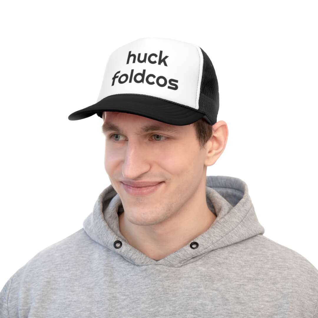 Man wearing a gray hoodie and a Foldcos Trucker Hat, which is black and white with huck Foldcos printed on the front.
