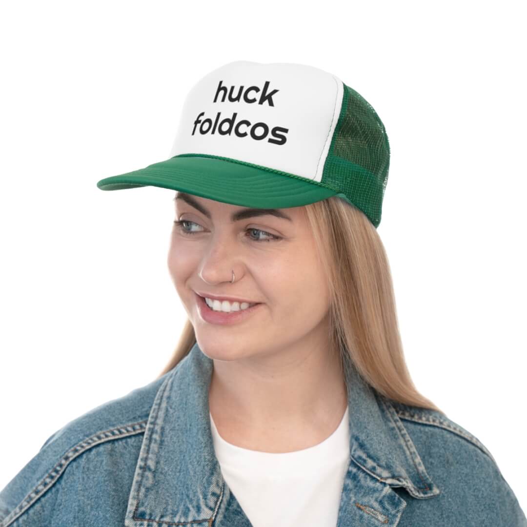 A smiling woman wears a green and white Foldcos Trucker Hat with the logo on front, pairing it with a denim jacket for a playful, casual look.