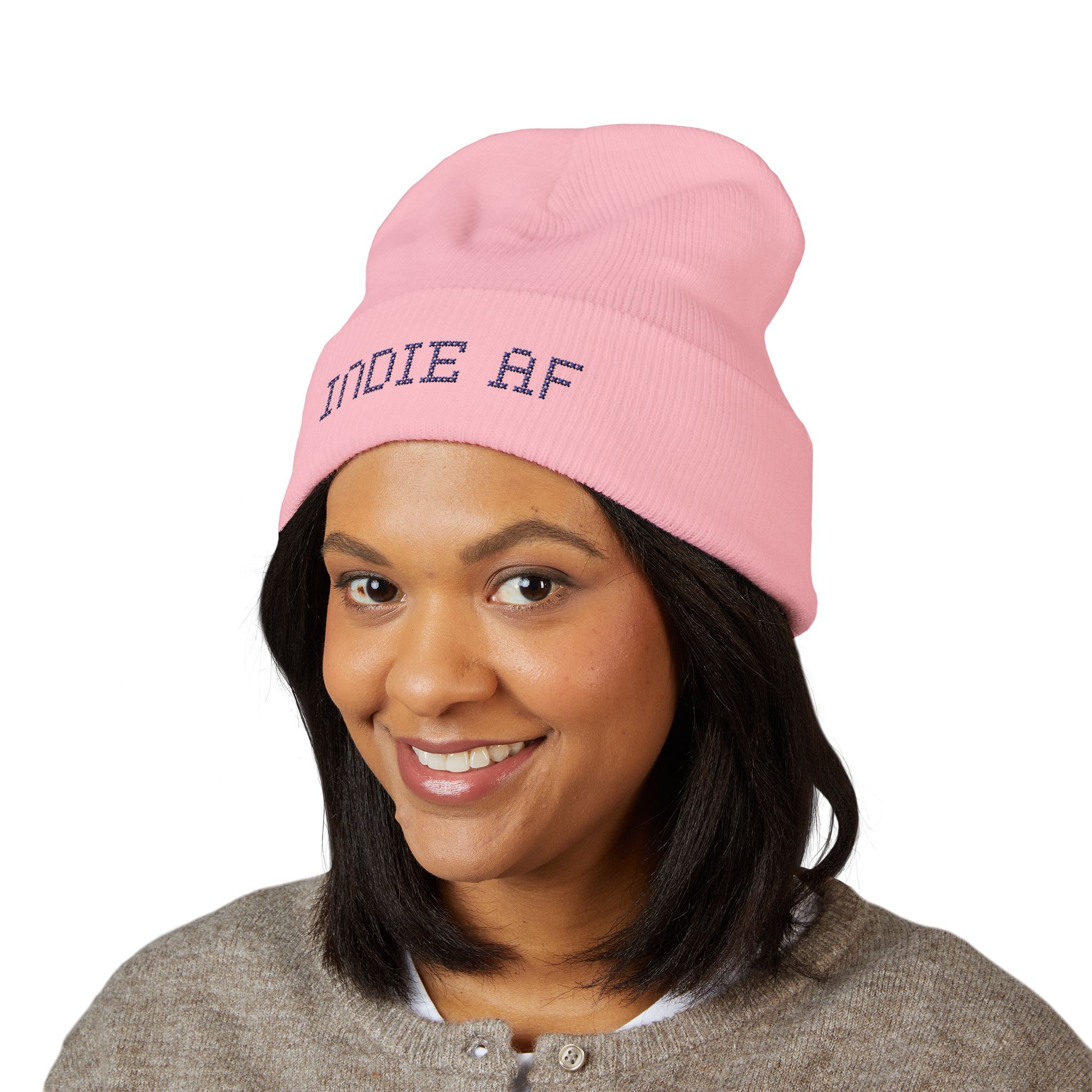 A woman smiles, wearing the Indie AF 2025 Holidays by Grown cuffed beanie in light pink with dark embroidery reading "INDIE AF," set against a plain white background.
